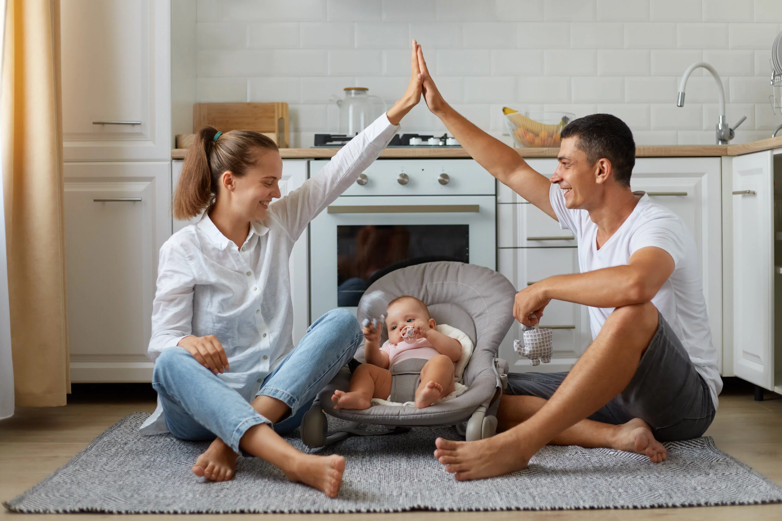 Full size photo of positive, three people mommy daddy small kid girl or boy in bouncer, parents make hands roof, enjoying sit on floor in light kitchen, indoors.